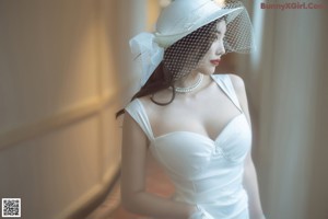 A woman in a white dress and hat leaning against a window.
